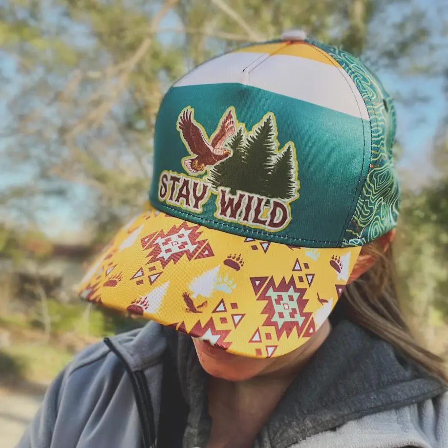 Woman wearing a hat that says Stay Wild with an eagle and pine trees. The hat bill is a vibrant yellow with an Aztec pattern and the rest of the hat is in a teal topo map print.